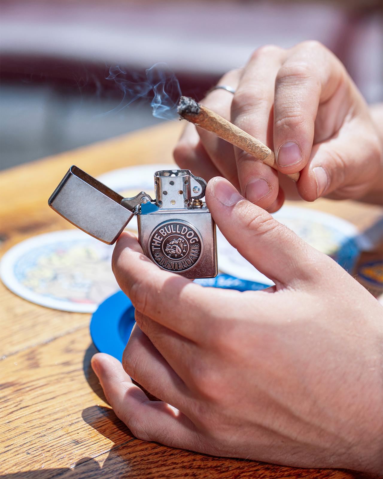 A person lighting a joint with a Bulldog-branded lighter at an outdoor table
